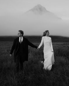 a man and woman holding hands walking through a field with a mountain in the background