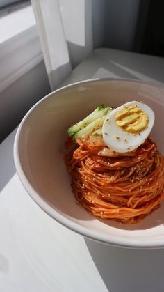 a white bowl filled with noodles and an egg on top of it, next to a window