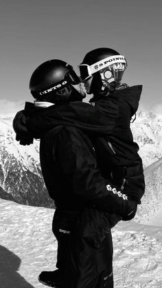 two snowboarders hugging each other on the top of a snowy hill with mountains in the background