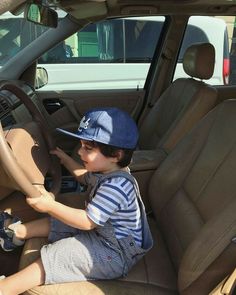 a young boy sitting in the driver's seat of a car wearing a hat