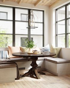 a wooden table sitting in front of two windows next to a bench with pillows on it