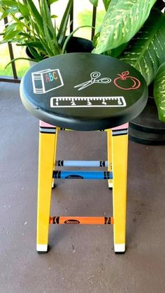 a stool that has been painted with different things on it and is sitting in front of some plants