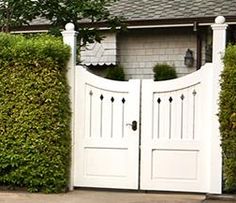 two white doors are open in front of a house with hedges on both sides and a brick wall behind them