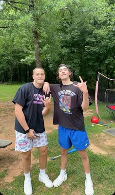 two young men standing next to each other in front of a net and trees, giving the peace sign