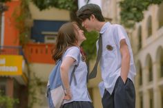 two young people standing next to each other in front of a building with trees and buildings behind them