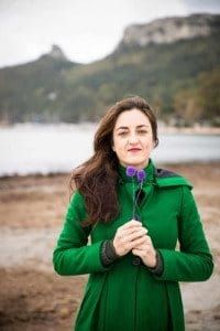 a woman in a green coat is holding a purple flower and looking at the camera