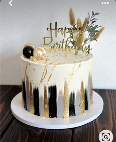 a white and black cake with gold lettering on it sitting on a wooden table next to a wall