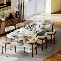 a dining room table set with white chairs and plates on it in front of a large window