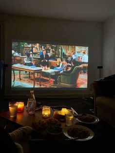 a couple of people sitting in front of a tv screen with food on the table