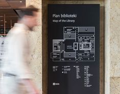 a man walking past a black sign on the side of a building that says plan biblioteiki map of the library