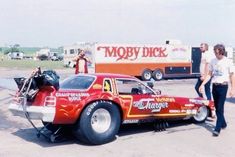 two people standing next to a red car with the hood up and an engine on it's flatbed