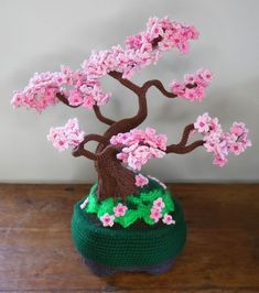 a crocheted bonsai tree with pink flowers in a green pot on a wooden table