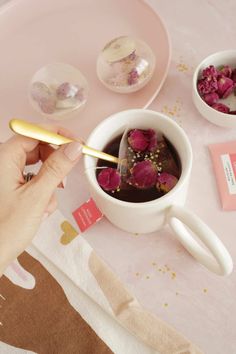 a person is holding a spoon over a cup filled with tea and pink flowers on a table