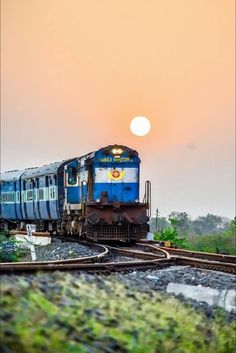 a blue and white train traveling down tracks under a cloudy sky with the sun in the background