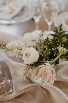 white flowers are in a glass vase on a table with wine glasses and napkins