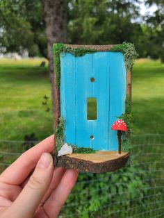 a hand holding up a miniature blue door with a mushroom on the outside and grass growing around it