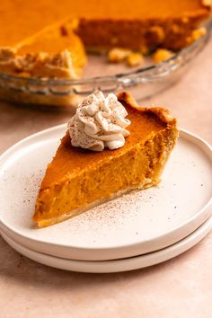 a slice of pumpkin pie on a white plate with whipped cream in the middle and a glass pie dish behind it