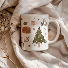 a white coffee mug with christmas trees on it sitting on a bed next to dried flowers