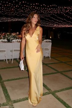 a woman in a long yellow dress standing next to a table with white chairs and lights