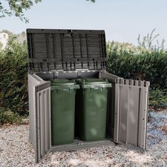 two green trash cans sitting in the middle of a gravel area next to some bushes