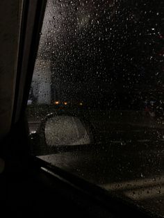 rain is falling on the windshield of a car in the dark, with street lights behind it