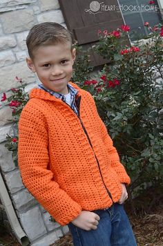 a young boy standing in front of a house wearing an orange sweater and jeans with his hands in his pockets