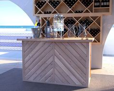 a wooden bar with wine glasses and bottles on it next to the ocean in front of an archway