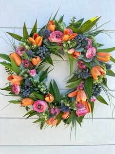 a wreath with orange and pink flowers is hanging on a white wooden wall next to greenery