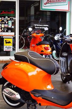 an orange scooter is parked in front of a store with several other mopeds