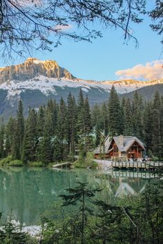 a cabin sits on the shore of a lake surrounded by pine trees and snow capped mountains