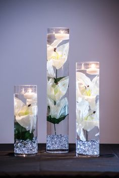 three clear vases with flowers in them on a table next to some water and candles