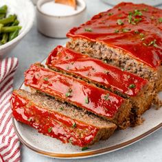 sliced meatloaf on a plate with sauce and green beans in the bowl next to it