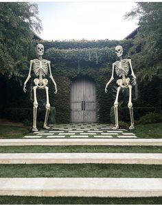 two skeleton statues standing in front of a door on some steps with grass and bushes