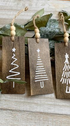 three wooden tags with white christmas trees on them hanging from twine strings next to greenery