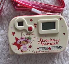 an electronic toy camera sitting on top of a white blanket next to a red bag