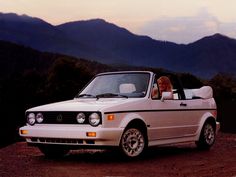 a woman sitting in the driver's seat of a white convertible car with mountains in the background