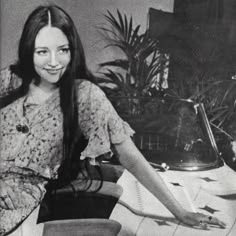 a woman sitting on top of a desk next to a chair and potted plant