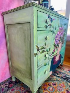 an old dresser painted green with flowers and butterflies on the drawers is sitting in front of a pink wall