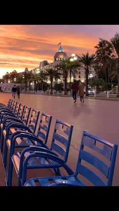 several blue chairs are lined up along the side walk in front of a building at sunset