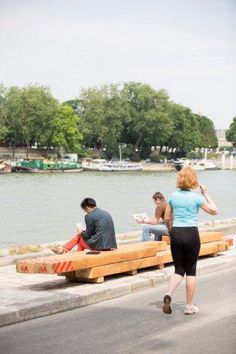 people are sitting on benches along the water's edge while one person walks by