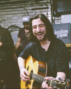 a man with long hair is playing an acoustic guitar and smiling at the camera while other men are standing behind him