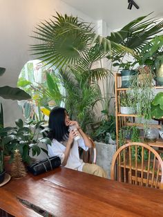 a woman sitting at a wooden table in front of potted plants and greenery