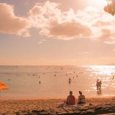 people are sitting on the beach under an umbrella and watching others play in the water