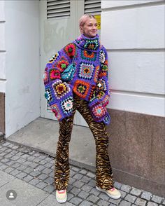 a woman standing in front of a white building wearing a colorful sweater and animal print pants