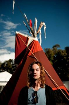 a man with long hair standing in front of a teepee