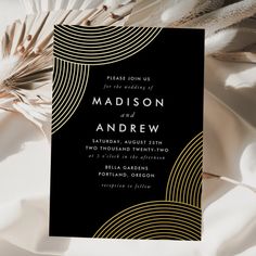 an elegant black and gold wedding card is on top of white fabric, with feathers in the background