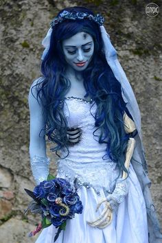 a corpse bride with blue hair and makeup holding a bouquet in her hand while standing next to a rock wall