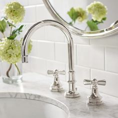 a bathroom sink with two faucets next to a mirror and flowers in a vase