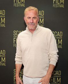 an older man standing in front of a black background wearing white pants and a beige sweater