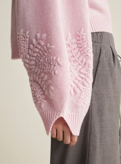 a woman wearing a pink sweater and grey pants with an embroidered flower on the side